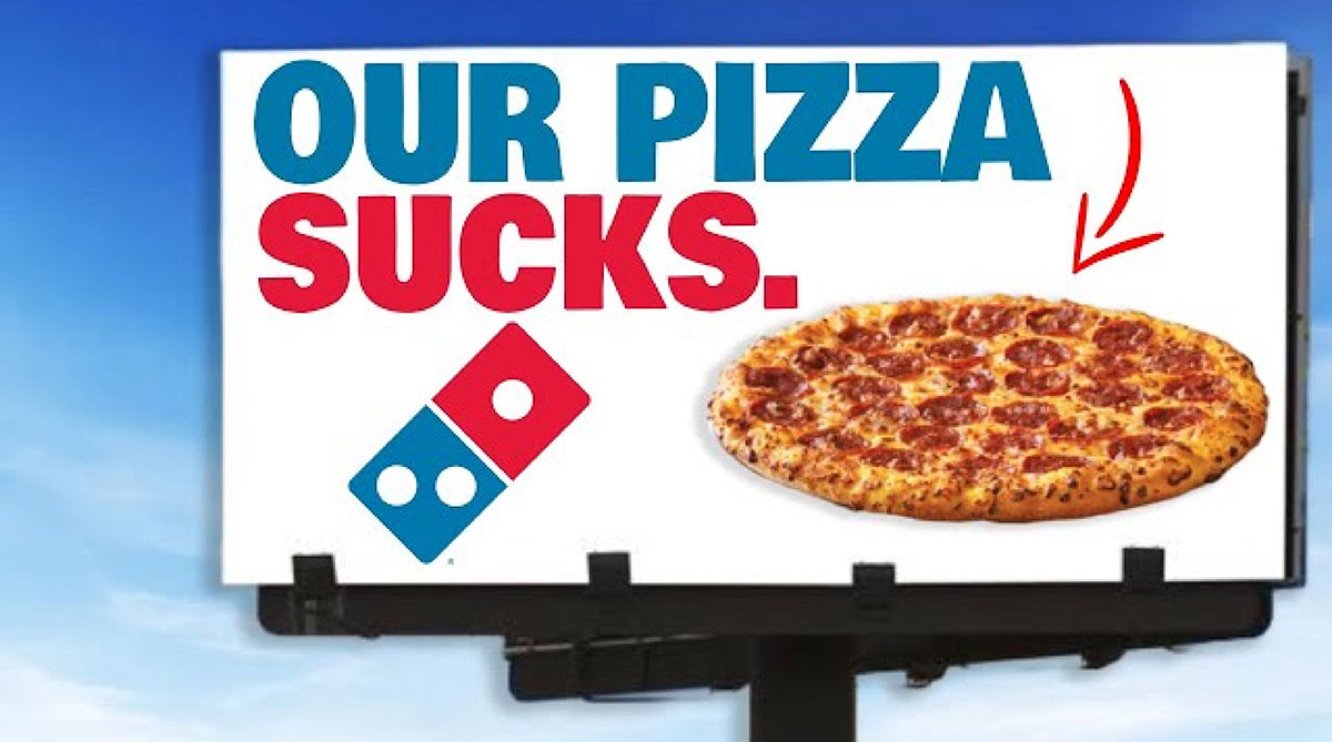 Domino's billboard with the headline Our Pizza Sucks, the Domino's logo and a pepperoni pizza under a blue sky